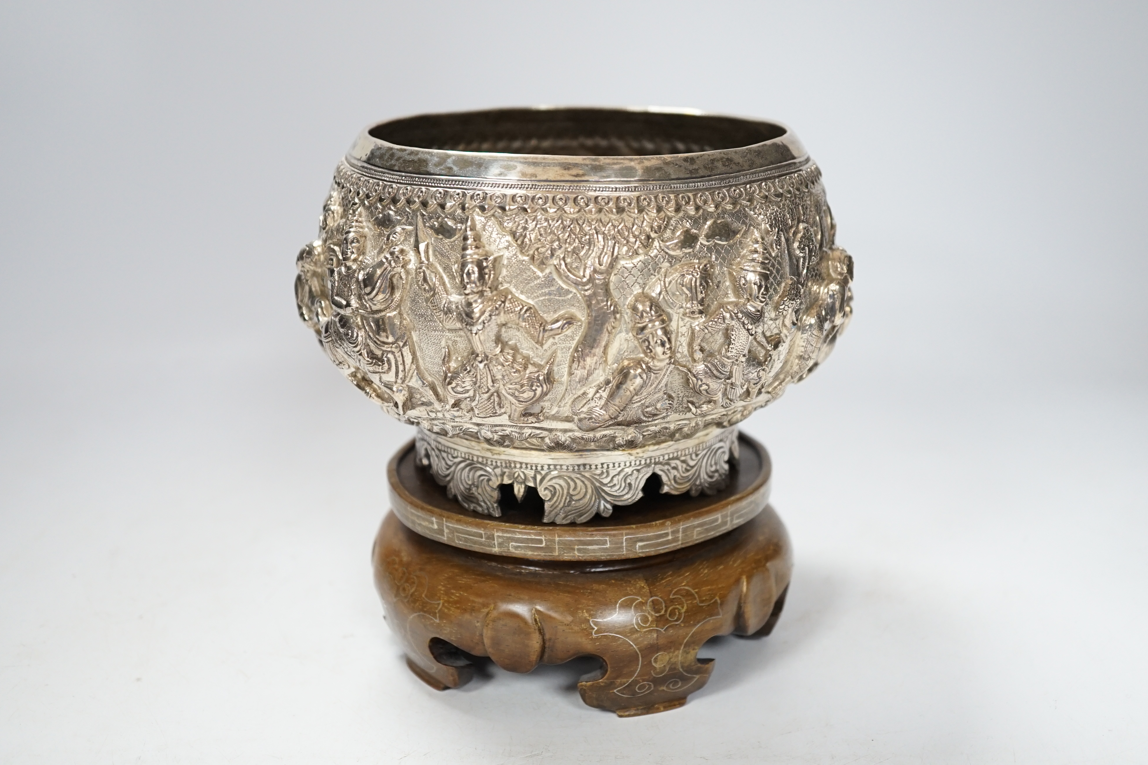 An Indian circular white metal bowl, decorated with a continuous band of figures in a landscape, on scrolled feet with hardwood stand, diameter 16cm